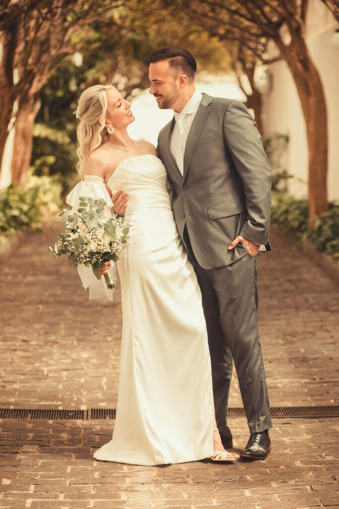 An elegant outdoor wedding scene showing a happy couple embracing under a tree-lined path.